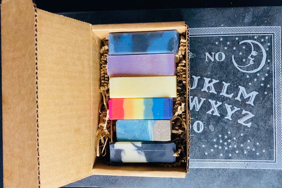 A cardboard box containing eight bar soaps of various colors, with each bar labeled with different color dyes used to create them.